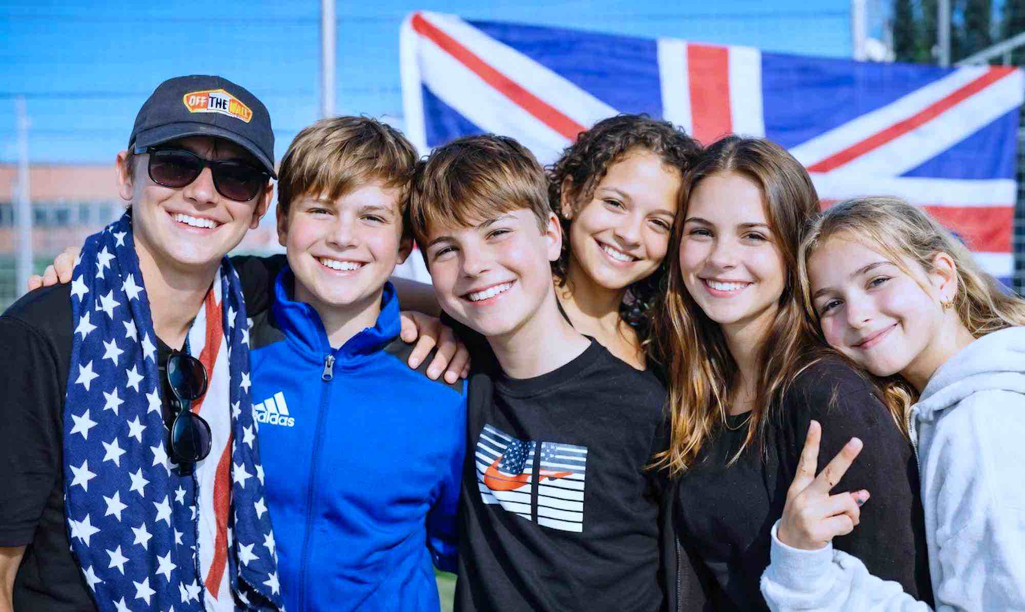 Jump Start English Project Weeks - Group of six happy teenagers smiling and posing together outdoors in front of a large Union Jack British flag. Fun English language project week photo.
