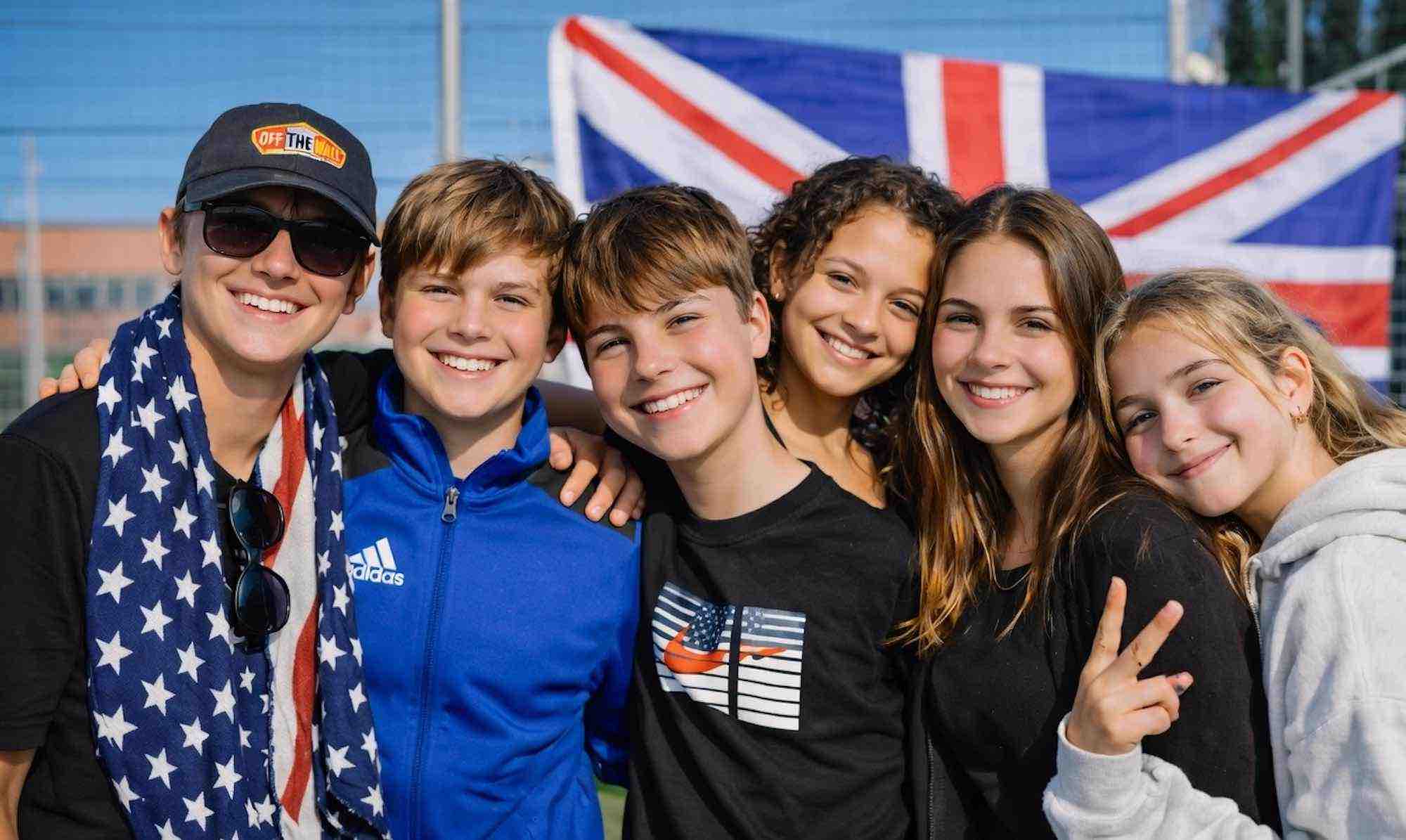Jump Start English Project Weeks - Group of six happy teenagers smiling and posing together outdoors in front of a large Union Jack British flag. Fun English language project week photo.